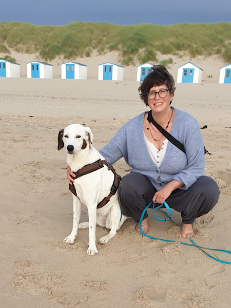 Frau hockt an einem Strand, neben ihr ein weißer Hund, den sie im Arm hält. Im Hintergrund sind Dünen und blau-weiße Strandhäuschen zu sehen.