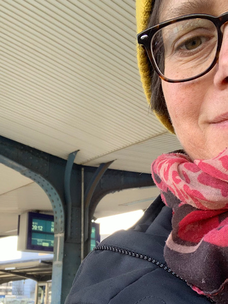 Rechts im Bild eine Frau mit gelber Mütze, dunklen Haare und Brille an einem Bahnhof stehend, im Hintergrund die Anzeigetafel und Bahnhofsarchitektur