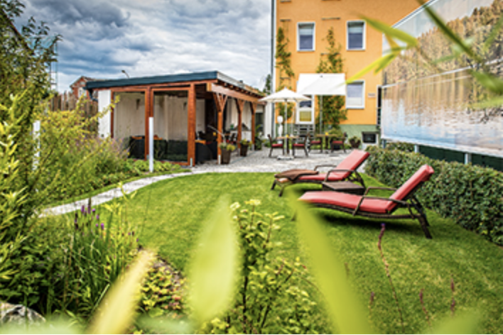 Zwei rote Liegestühle auf einer Wise, vor einem überdachten Außenbereich und einem gekben Haus im Hintergrund