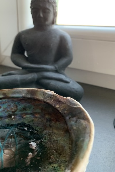 dunkelgraue kleine Buddhafigur, davor eine Muschel mit einem salbeiräucherbündel