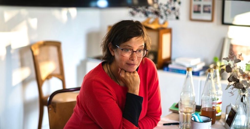 Frau sitz am Tisch, stützt ihren Kopf in die Hand und blickt aufmerksam und zuhörend ihr Gegenüber an.