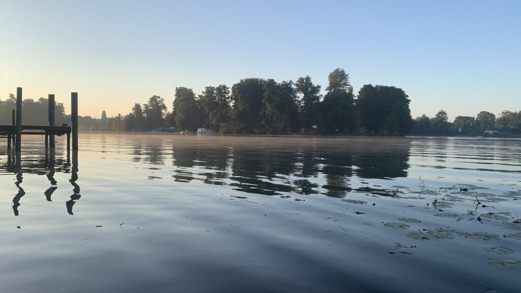 Blick über einen See, mit Morgennebel. Im Hintergrund Bäum und davor ankernde Schiffe