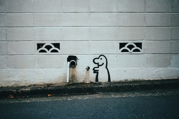 eine weiße Wand mit einem offenen Abflussrohr. An der Wand ist in schwarzen Kontirlinien eine lachende Figur gezeichnet. Im Vordergrund ist die Straße noch erkennbar.