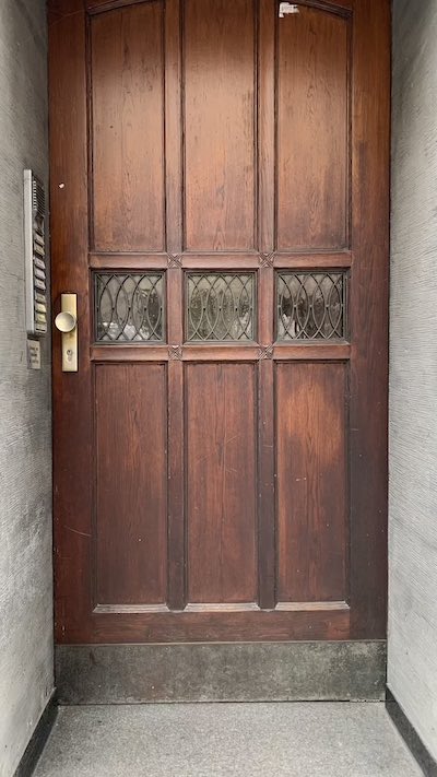 Eine alte Eingangstür eines Wohnhauses aus dunklem Holz mit einem goldenen Knauf und drei kleinen Fenstern in der Mitte der Tür.