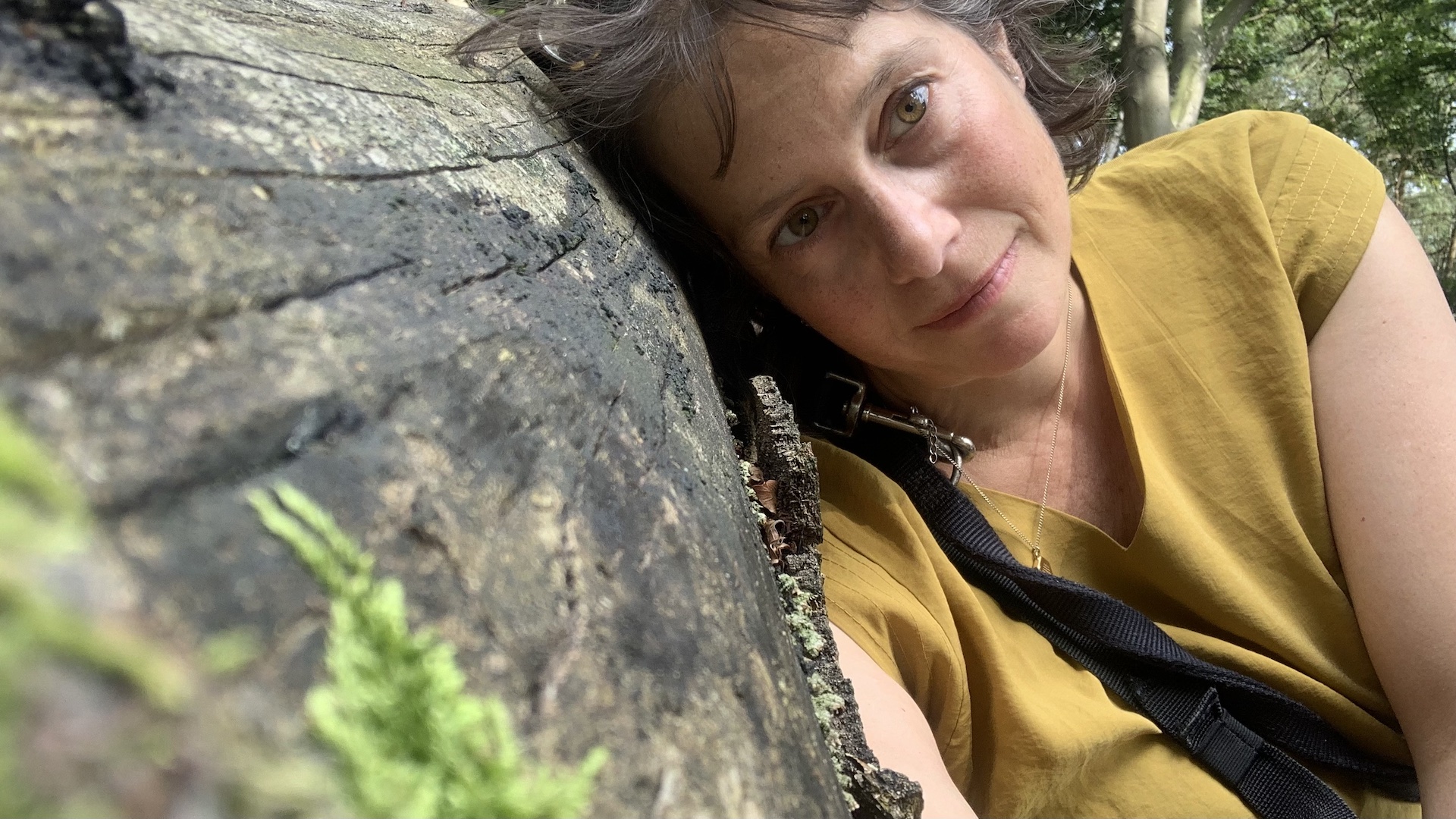 Frau lehnt sitzen dann einem Baumstamm und blickt verträumt in die Kamera.