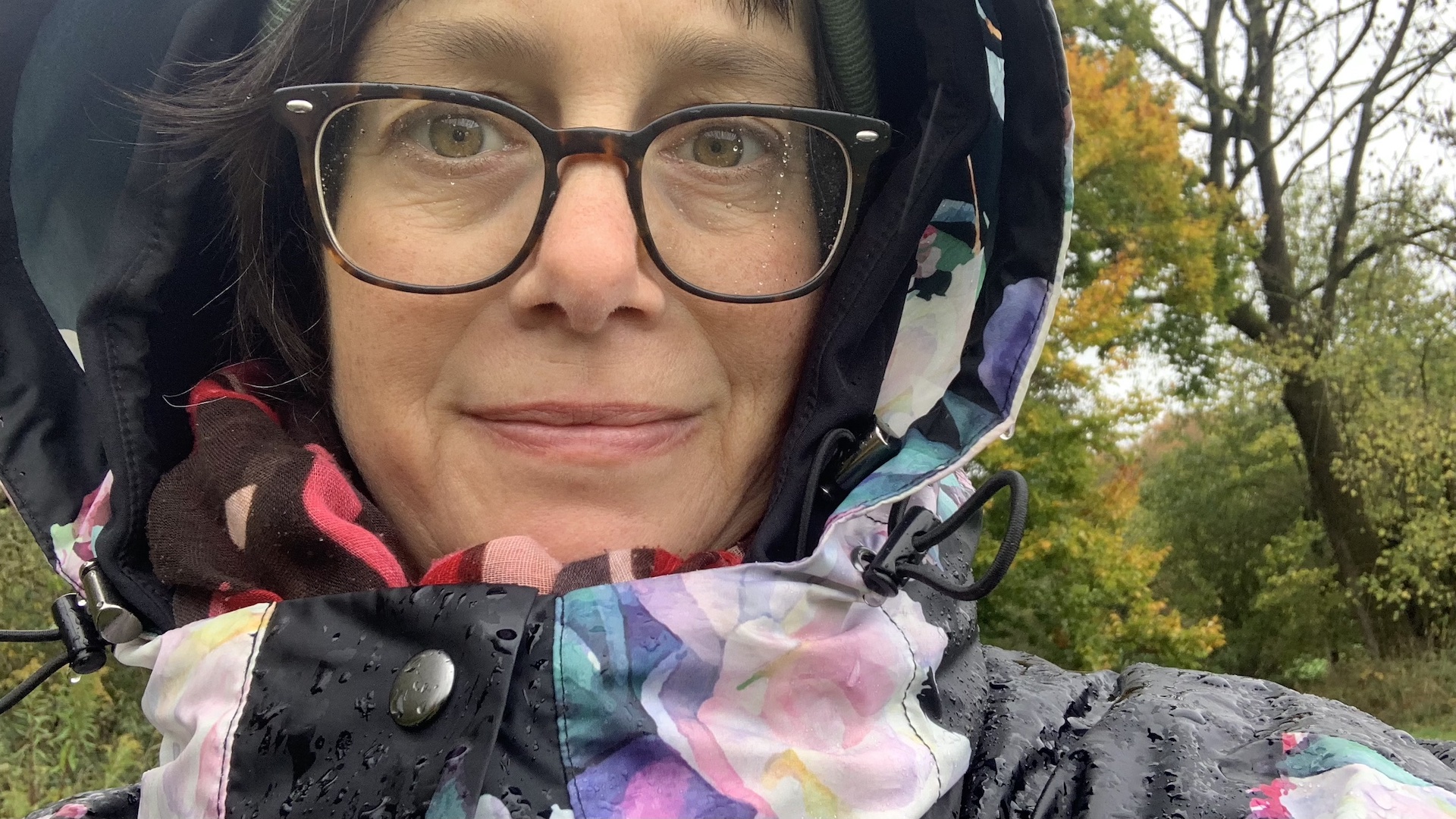 Selfie Katja Reetz mit Kapuze und buntem Regencape, auf der Brille Regentropfen