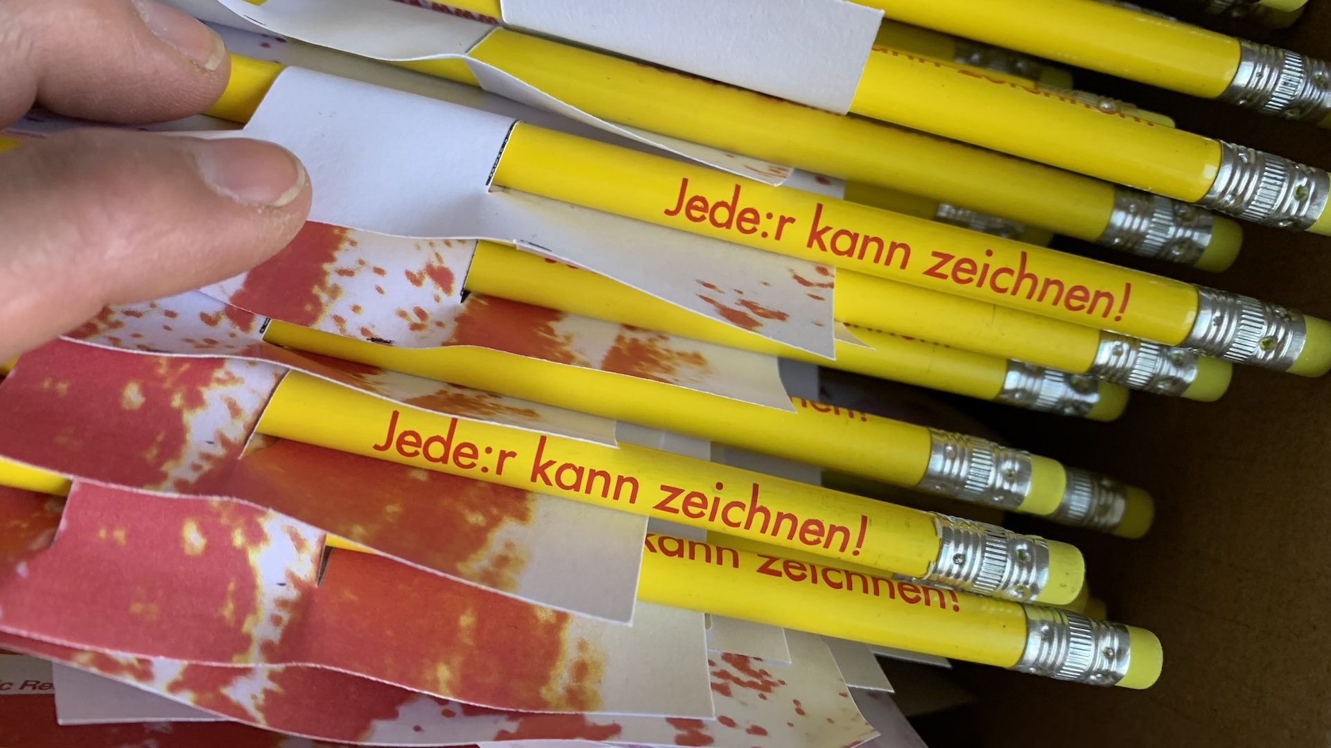 Handvoll gelber Bleistifte mit dem roten Aufdruck: "Jede:r kann zeichnen!"