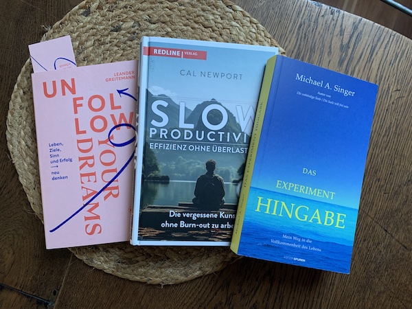 Drei Bücher, von oben fotografiert. Unfollow your Dreams (roter Schriftzug auf rosa Cover), Slow Productivity (Motiv = Meditierender Mann vor Berglandschaft), Experiment Hingabe (gelber Text auf blauem Meermotiv)