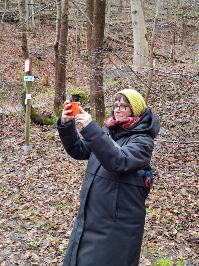 Porträt im Wald: Eine Frau fängt mit ihrem Smartphone ein Detail ein und lässt gleichzeitig den Blick zur Seite schweifen, um neue Perspektiven für das 2. Quartal 2026 zu entdecken. Das Bild unterstreicht die Verbindung von Fokus und strategischer Weitsicht.