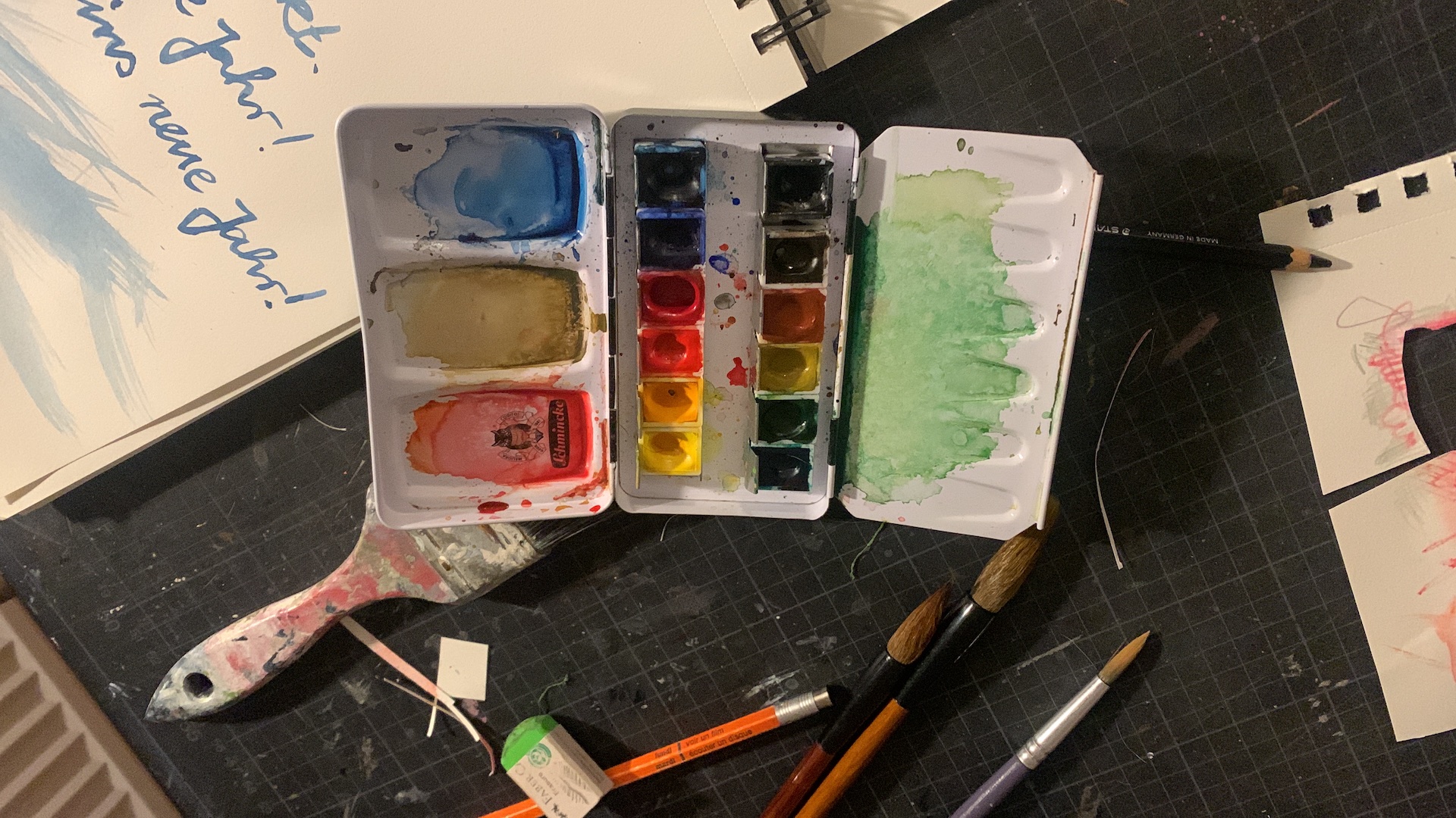 Draufsicht auf einen Aquarell-Arbeitsplatz mit Farbkasten, Pinseln, Bleistift, Radiergummi und einem Skizzenbuch. Das Skizzenbuch zeigt strukturierte Diagramme und klaren, handgezeichneten deutschen Text: "105 FAKTEN", "PROZESSE STEUERN", "HANDLUNGSFÄHIG WERDEN". Auf der Schneidematte liegen visuelle Anker wie "PROZESS-ENERGIE" und "FOKUS". Das Bild vermittelt einen organischen, aber strukturierten Prozess-Strukturierungsansatz durch visuelle Mittel.