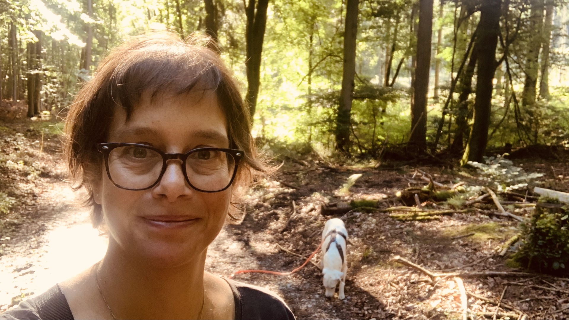 Porträt Katja Reetz im spätsommerlichen, sonnigen Wald. Ein weißer Hund schnüffelt am Waldboden.