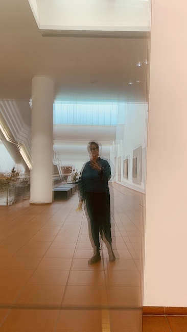 Aufnahme einer Person vor einer Spiegelwand im Museum Ludwig, Köln. Das Bild wird durch die Staffelung der Glasscheiben verschwommen. 